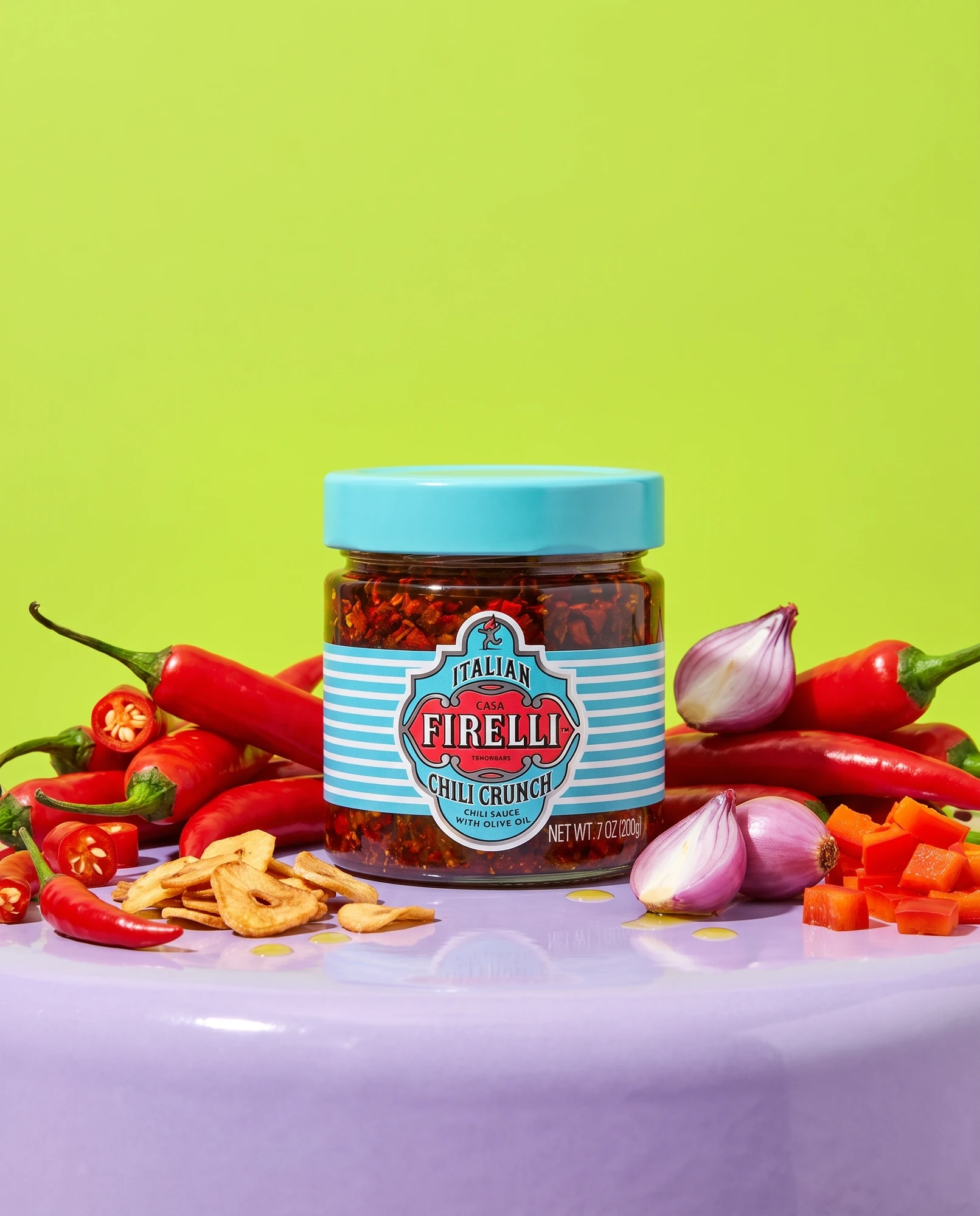 Jar of Casa Firelli Chili Crunch among fresh chillies, garlic chips and onion against a lime-green background