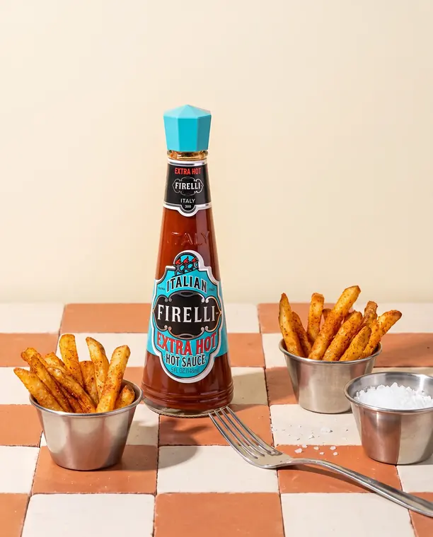 Bottiglia di Extra Hot Sauce Casa Firelli con patatine speziate su piano a scacchi color terracotta