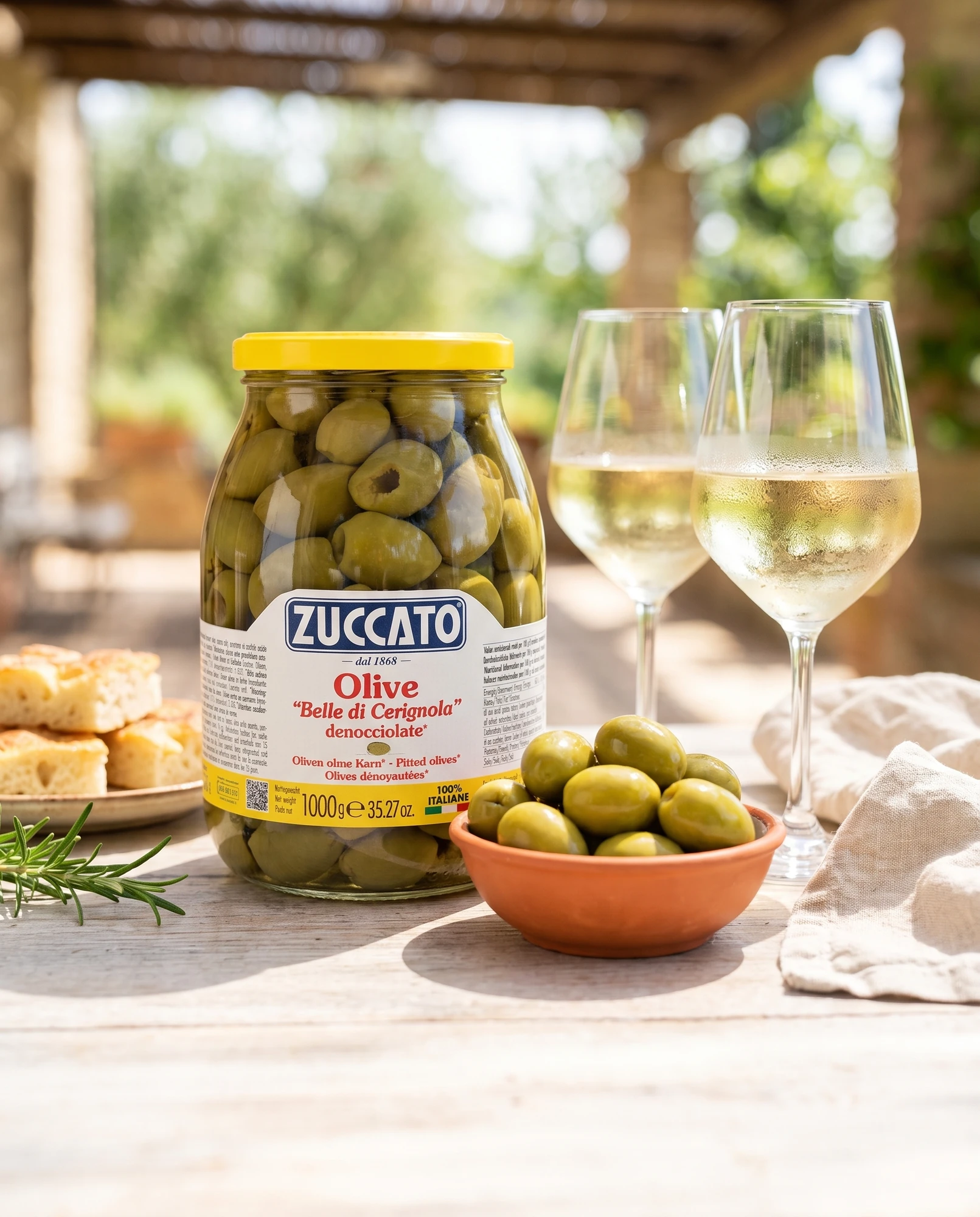 Jar of Zuccato pitted Belle di Cerignola olives on an outdoor table with glasses of white wine