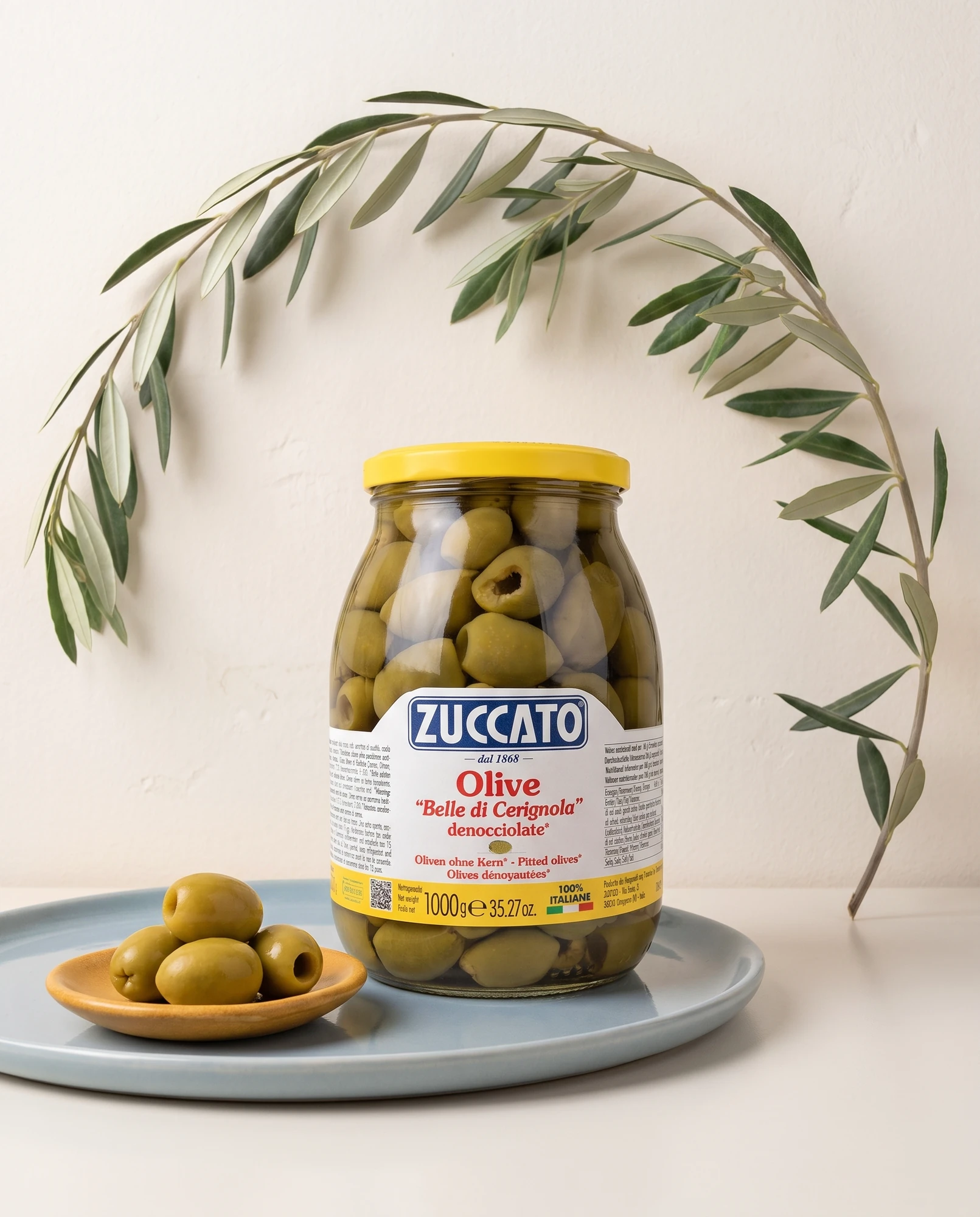 Jar of Zuccato Belle di Cerignola olives with an olive branch and a bowl of olives on a blue plate